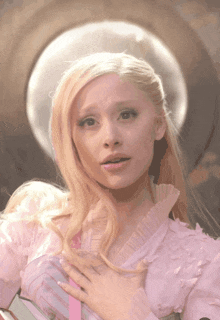 a blonde woman in a pink dress holds her hand to her heart