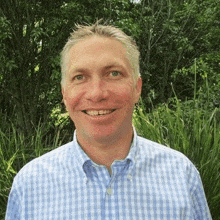 a man in a blue and white plaid shirt is smiling in front of a forest .