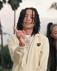 a young man with dreadlocks is wearing a yellow jacket with a heart on it and clapping his hands .