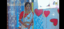 a bride in a white saree is standing in front of a wall with red hearts .