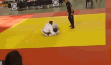 a referee is standing on a yellow mat watching a judo match between two fighters .