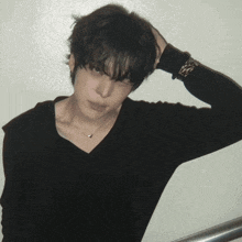 a young man wearing a black sweater and a necklace holds his hand to his head