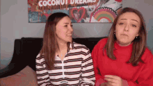 two women are sitting on a couch in front of a painting that says coconut dreams