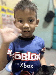 a young boy wearing a blue roblox shirt holds a cell phone