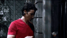 a man in a red shirt is standing in front of a wall and looking at his phone .