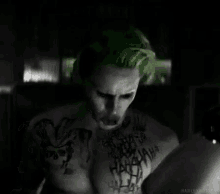 a man with green hair and a joker tattoo on his chest is crying in a dark room .