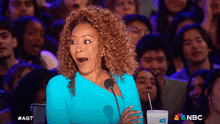 a woman with a surprised look on her face is sitting in front of a crowd with a nbc logo on the bottom right