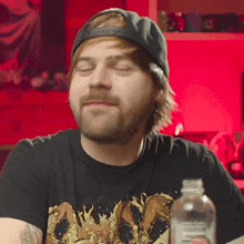 a man wearing a baseball cap and a black shirt with a dragon on it is sitting in front of a bottle of water