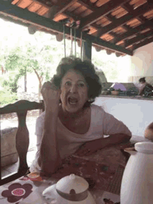 an elderly woman is sitting at a table with her mouth open and making a funny face .