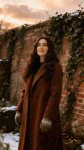 a woman in a brown coat and gloves is standing in front of a brick wall