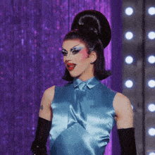 a drag queen wearing a blue top and black gloves stands in front of a purple curtain