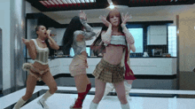 three women are dancing in a kitchen wearing skirts and crop tops .