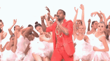 a man in a red suit is singing into a microphone surrounded by white ballerinas