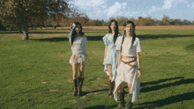 three women are standing in a grassy field wearing white dresses