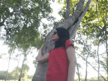 a woman in a red shirt leans on a tree