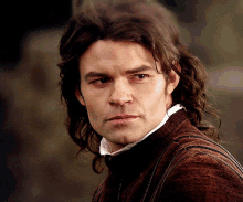 a man with long hair is wearing a brown sweater and white shirt