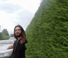 a man with long hair and glasses stands in front of a hedge with a license plate that says 8 be