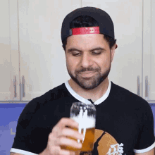 a man with a beard wearing a black shirt and a baseball cap is drinking from a glass of beer