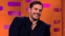 a man in a suit and black shirt is smiling while sitting in front of an orange background .