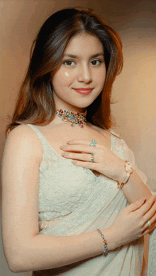 a woman wearing a necklace and a bracelet stands with her hands on her chest