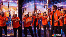 a group of men in orange shirts are performing on a stage with a nbc logo in the background
