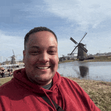 a man in a red hoodie is smiling in front of a lake with windmills in the background