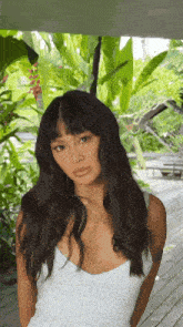 a woman with long dark hair and bangs is wearing a white dress and standing on a wooden deck .