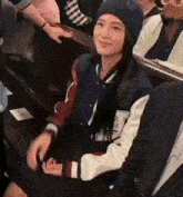 a woman wearing a baseball jacket and a beanie is sitting in a crowd of people .
