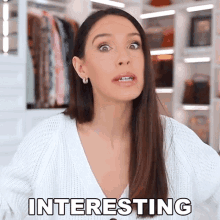 a woman in a white sweater is standing in front of a closet and the word interesting is on her face .