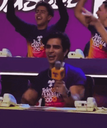 a man wearing a teleto camb t-shirt is sitting at a table