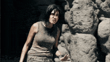 a woman in a tank top is standing in front of a wall of rocks