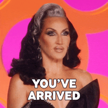 a drag queen says " you 've arrived " on a pink and orange background
