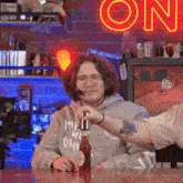 a man sitting at a table with a bottle of beer and a neon sign in the background that says one