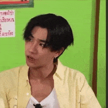 a young man in a yellow shirt is making a funny face while sitting in front of a green wall .