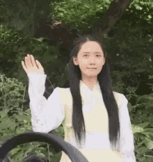 a woman with long black hair is standing next to a steering wheel and waving her hand .