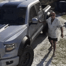 a man is standing in front of a silver truck on a dirt road