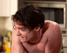 a shirtless man wearing a necklace is standing in a kitchen with his head down .