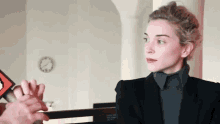 a woman wearing a black jacket and a grey turtleneck is sitting in front of a clock on the wall