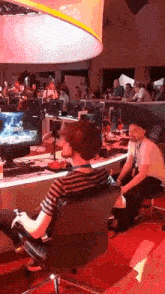 a man sits in a chair playing a video game while another man sits at a desk playing a video game