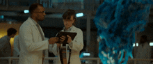 two men in lab coats are looking at a tablet and a blue object