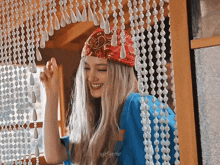 a woman wearing a bandana and a blue shirt smiles in front of a beaded curtain