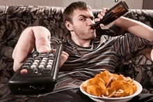 a man sits on a couch drinking beer and holding a remote control