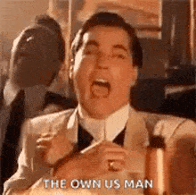 a man in a suit and tie is shouting the own us man while holding a glass of wine .