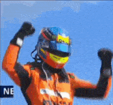 a man wearing a helmet and an orange jacket is raising his arms in the air .