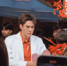 a man in an orange shirt and a white jacket stands in front of flowers