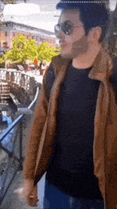 a man wearing sunglasses and a brown jacket is standing in front of a building .