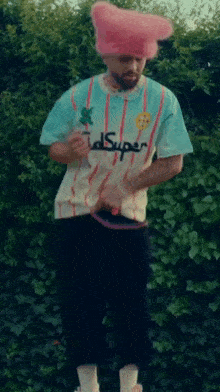 a man wearing a pink hat and a shirt that says super is standing in front of a bush .