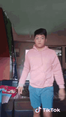 a young man in a pink hoodie and blue shorts is dancing in a living room with tiktok written on the bottom right