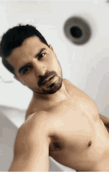 a shirtless man with a beard takes a selfie in front of a white wall