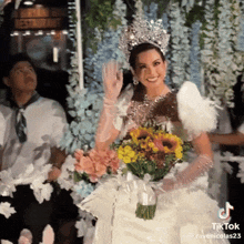 a woman in a wedding dress holds a bouquet of flowers and a crown on her head